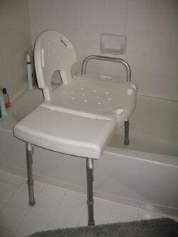 A white plastic bench sits with two legs inside a bathtub and two legs outside the tub resting on the floor.