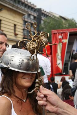 smiling woman wearing a colander on her head being "blessed" by a brass Flying Spaghetti Monster in the style of a Roman Catholic scepter.