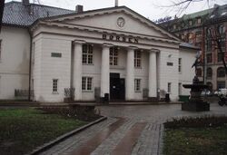 Photograph showing the main entrance to the Oslo Børs, known in English as the Oslo Stock Exchange in Norway. The building is in the Renaissance architectural style, its exterior painted in an Ivory white colour. Its entrance is protected by an imposing porch topped with a triangular pediment supported by four columns.