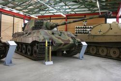 A three quarters view of a large tank with a flat-faced turret, dull yellow, green and brown wavy camouflage, on display inside a museum. The frontal armour is sloped. The long gun overhangs the bow by several meters. Two waist-high cartridges sit on their bases in front of it.