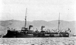 A large, black ship lays off shore; heavy canvas awnings have been erected on the deck to shelter the crew.