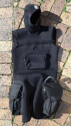 Sleeveless tunic in thin but abrasion resistant neoprene, with integral hood, two cargo pochets on the sides of the thighs, cross-chest zip closure, front pocket on the torso and opening for access to a dry-suit inflation valve. The tunic is suitable for wearing over most one-piece wetsuits for extra insulation, but mainly to support the cargo pockets and hood.