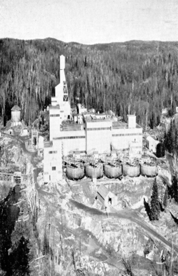 Boxy buildings and the mine's head shaft behind ten large tanks, surrounded by trees