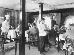 A black-and-white photograph of an interior scene. Four men and three women in semi-formal dress are seated on dining chairs decorated in a floral motif around two small square tables. Two staff wearing white are serving them food and drinks. The one on the left is wearing a chef's hat. A man wearing dark clothes is looking towards the camera as he walks across the room. There are two substantial square-profile vertical columns in the room, three dome-type electric lights are visible in the ceiling, and there are two large windows behind, with elaborate curtains. The walls are covered in striped and patterned wallpaper.