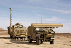 A Mastiff fitted with Choker Mine Rollers following on behind the Panama remote control Land Rover. The vehicles are in a flat desert in Jordan with a bright blue sky. At the front of the Land Rover, about six feet off the ground, is a horizontal beam which can be lowered in front of the vehicle to scan the ground with radar. The beam is approximately three metres wide.