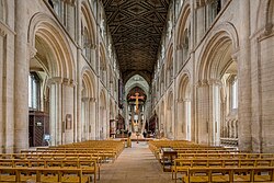 This interior view of Peterborough shows part of the very long nave and the chancel with no screen dividing the building. The structure is mostly of a unified Norman appearance with three levels of simple round-topped arches, only the tall arches of the central tower being Gothic. Other visible features are the ancient painted ceiling, the medieval choir stalls and the modern Rood, which has a gilt wooden figure of Christ on a red cross suspended high in front of the tower arch.