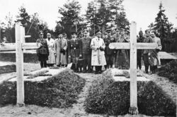 17 men, most in military uniform, stand in a cemetery, inspecting two graves.