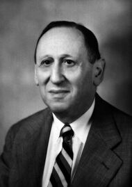 Balding man in his early 60s in coat and tie, with a serious but slightly smiling expression