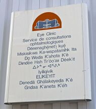 A metallic white sign on a gray background with a red and blue depiction of a building at the top. Below it is text in blue saying "Eye Clinic" in English, French and the other nine official territorial languages