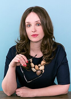A woman sitting at a desk, with pale skin and dark wavy hair; she is wearing a seashell necklace and holding a pair of glasses