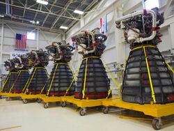 Six rocket engines, consisting of a large bell-shaped nozzle with working parts mounted to the top, stored in a large warehouse with white walls decorated with flags. Each engine has several pieces of red protective equipment attached to it and is mounted on a yellow wheeled pallet-like structure.