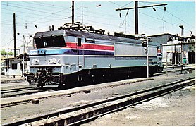 A gray electric locomotive with a black roof and red and blue side stripes