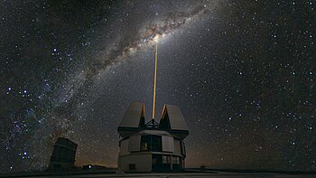 In the foreground, an astronomical observatory emits a ray of light vertically. In the background is a cluster of stars in the night.