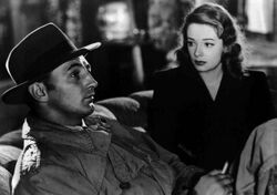 Black-and-white image of a man and a woman sitting side by side on a couch, viewed at an angle. The man, in profile in the left foreground, stares off to the right of frame. He wears a trenchcoat, and his face is shadowed by a fedora. He holds a cigarette in his left hand. The woman, to the right and rear, stares at him. She wears a dark dress and lipstick of a deeply saturated hue.