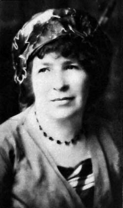 Portrait photo of Catherine Amy Dawson Scott. She is pale with dark darly hair that falls to her chin, a slight smile on her face as she gazes off toward the left, and is wearing an ornate flower hat, beaded necklace, and possibly a cardigan over a dress.