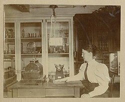 Sitting in the Wellesley physics laboratory, Chase places her hand on a glass photographic plate on a table below a Crookes tube, to take a radiograph of the bones in her hand.