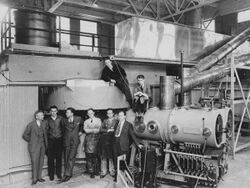 Six men in suits and ties stand in front of gigantic machinery. Two more are sitting on top of it.