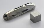A bar and a cube of a silvery metal crystal lying on a grey surface