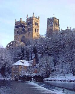 A winter view showing Durham Cathedral with three large towers looming high on a craggy cliff above a river bordered with snow-covered trees, a weir and a house.
