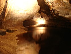 Cave passage with with dark water flowing towards the viewer beside a silty bank on the left. The cross-section of the passage is roughly triangular, with a flat, wide bottom, and a relatively smooth slanting roof on either side.