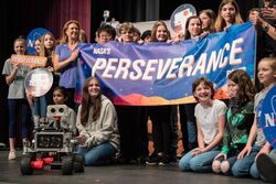 About twenty K-12 student finalists are standing on a stage, all smiling, and holding a banner that reads "NASA's perseverance rover". In front of them on the stage is a miniature rover.