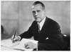 A photographic portrait of a man wearing a suit seated at a desk with a pen in his hand