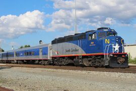 A passenger train with a diesel locomotive. The cars are grey with a blue window stripe and a red sill stripe. The locomotive is blue and gray with a black roof and red sill stripe.