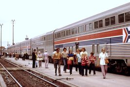 A train of two-level passenger rail cars at a station. The coaches are stainless steel, with red and blue horizontal stripes halfway up their sides