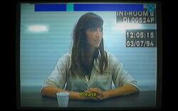 Old computer footage of a 37-year-old woman in a white shirt with long brown hair, sitting at a police interview desk. She is holding her hands together in front of her, and is looking to the right of the camera. There is a subtitle that reads "Please." At the top of the screen is a scroll bar, positioned at one-third.