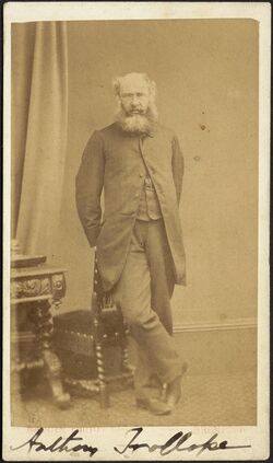 Photograph of Anthony Trollope leaning against a chair, his right leg crossed over the left. His name is written in ink at the base of the image.