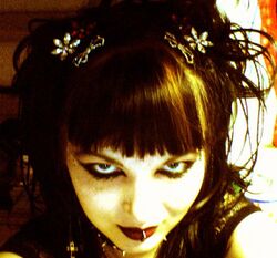 Very contrasted photograph of the head of a young white woman looking up at the viewer. She smiles. She wears goth makeup. She has a fringe.