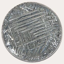 A shiny silver-white medallion with a striated surface, irregular around the outside, with a square spiral-like pattern in the middle