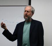 A gray-haired man with a gray beard wearing a black jacket and gray shirt, talking with a whiteboard behind him.