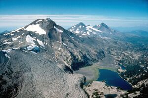 Aerial view from the southeast looking north of the Three Sisters volcanoes, three mountains sparsely covered with ice and snow. From left to right the image shows South Sister, Middle Sister, and North Sister, with a black lava flow in the left foreground.