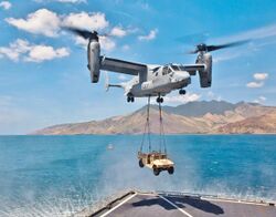 An MV-22 Osprey with its rotors up to vertical with a HMMWV vehicle hanging by two sling wires.