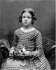 Three-quarter length studio photo of seated girl about nine years old, looking slightly plump and rather solemn, in a striped dress, holding a basket of flowers on her lap