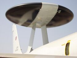 Close-up view of black disc-shaped radar with wide diagonal white band. The radar rests on 2 convergent struts above aircraft fuselage.