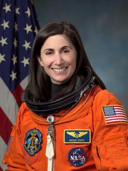 An astronaut wearing her orange mission suit with the American flag embroidered on her shoulder and name tag and mission patches embroidered on her chest.