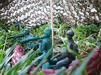 Three green plastic bazooka men on dirt in between several strands of grass