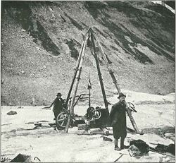 A man standing on a glacier with a drilling rig and a mountain ridge behind him.
