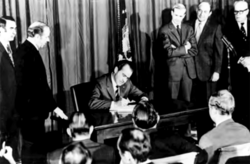Photograph showing President Nixon seated at a desk signing the bill, with multiple people watching him; those to his right and left are standing, and those facing him in the foreground are seated