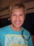 A man in a light blue t-shirt smiles towards the camera. He has blond tousled hair, and sports a stubble.
