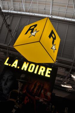 A large orange box with the letter "R" and a star hangs from a tall building. Below it is a convention booth with the lit-up text "L.A. NOIRE". The game's cover art is vaguely seen beneath the logo.