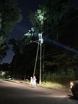 Two utility linemen with hard hats work to replace a cutout fuse 35 feet above them with a non-conductive pole with a hook on the top.