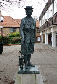 Full-length statue on a raised plinth of Sayers standing, a cat walking at her feet