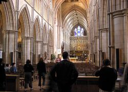 This longshot of the interior of Southwark Cathedral shows both the nave, with crisply detailed pointed arches, and the older structure beyond. A number of visitors are silhouetted against the pale stone.