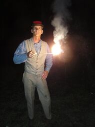 A man is standing in the dark. He is holding out a short stick at mid-chest level. The end of the stick is alight, burning very brightly, and emitting smoke.