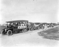 A black and white photograph of a line of at least seven open-air buses filled with potential real estate investors, showing banners that read "HI-A-LE-AH", stopped on white dirt roads surrounded by lawns in undeveloped neighborhoods; some houses in the background