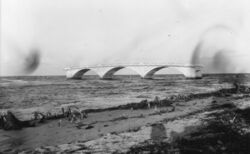 A black and white photograph of the ruins of a bridge taken from a beach with broken and uprooted trees recently damaged by a hurricane