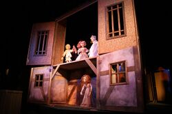 A woman performs a puppet show by manipulating puppets that are situated on a stage, stylized as a wooden playhouse.